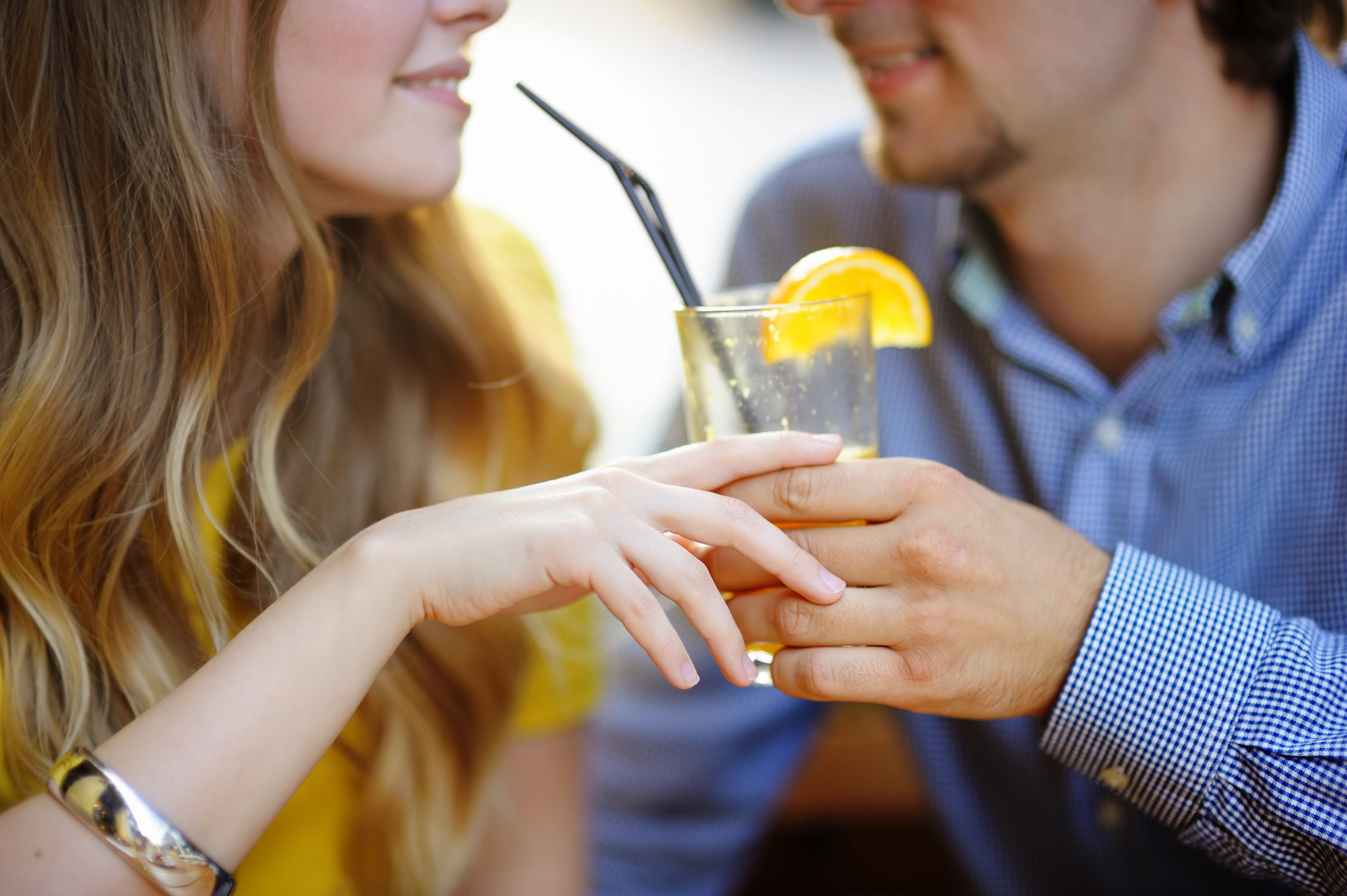 A close-up of a couple holding a glass with an orange slice garnish and a straw. The woman has long hair, wearing a yellow top, and the man has short hair, wearing a blue checkered shirt. They are smiling and looking at each other.