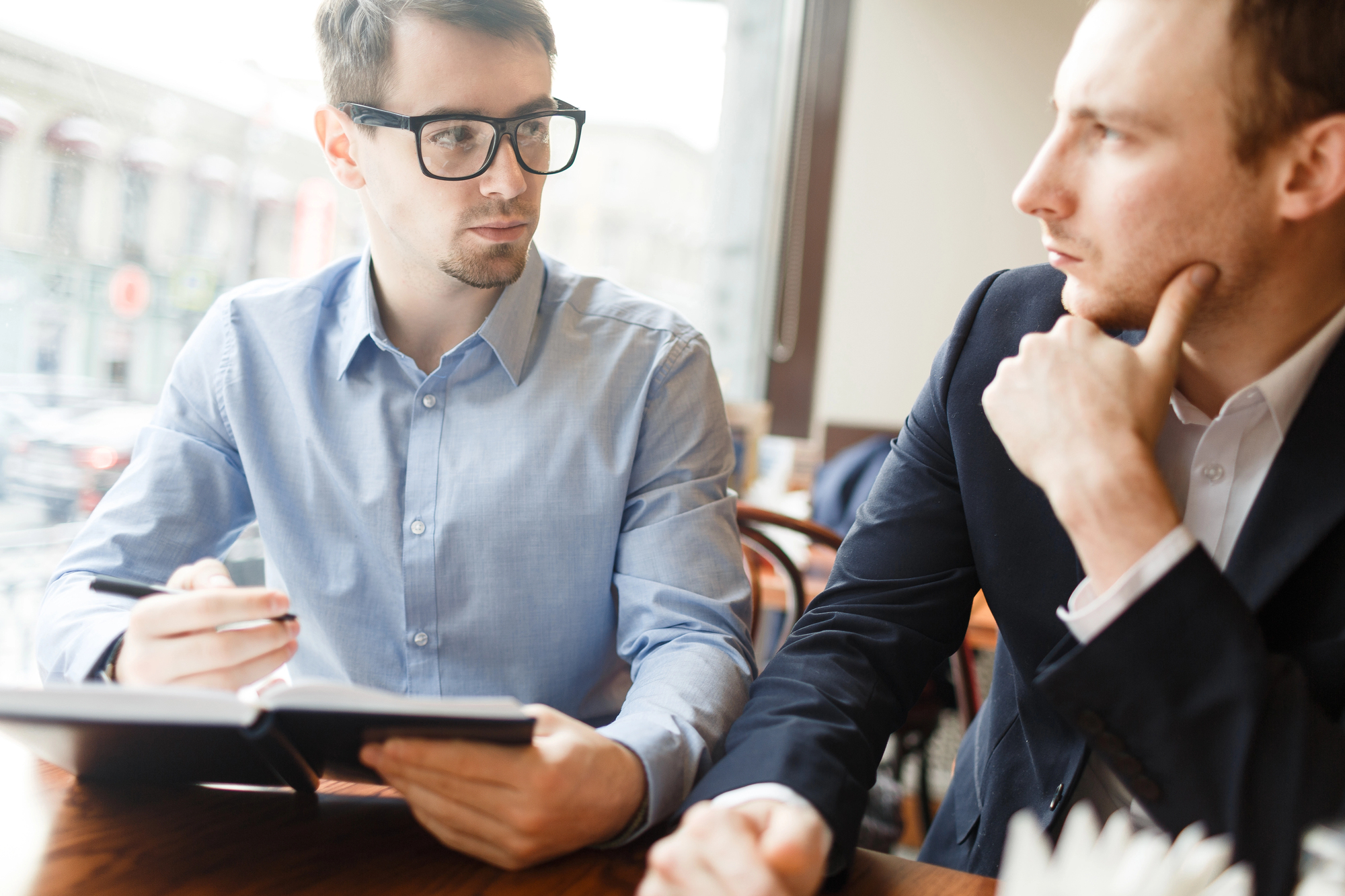 Two men sitting at a table in a cafe, engaged in a conversation. One is wearing glasses and holding a notebook, while the other has his hand on his chin, listening attentively. Light pours in through a large window behind them.