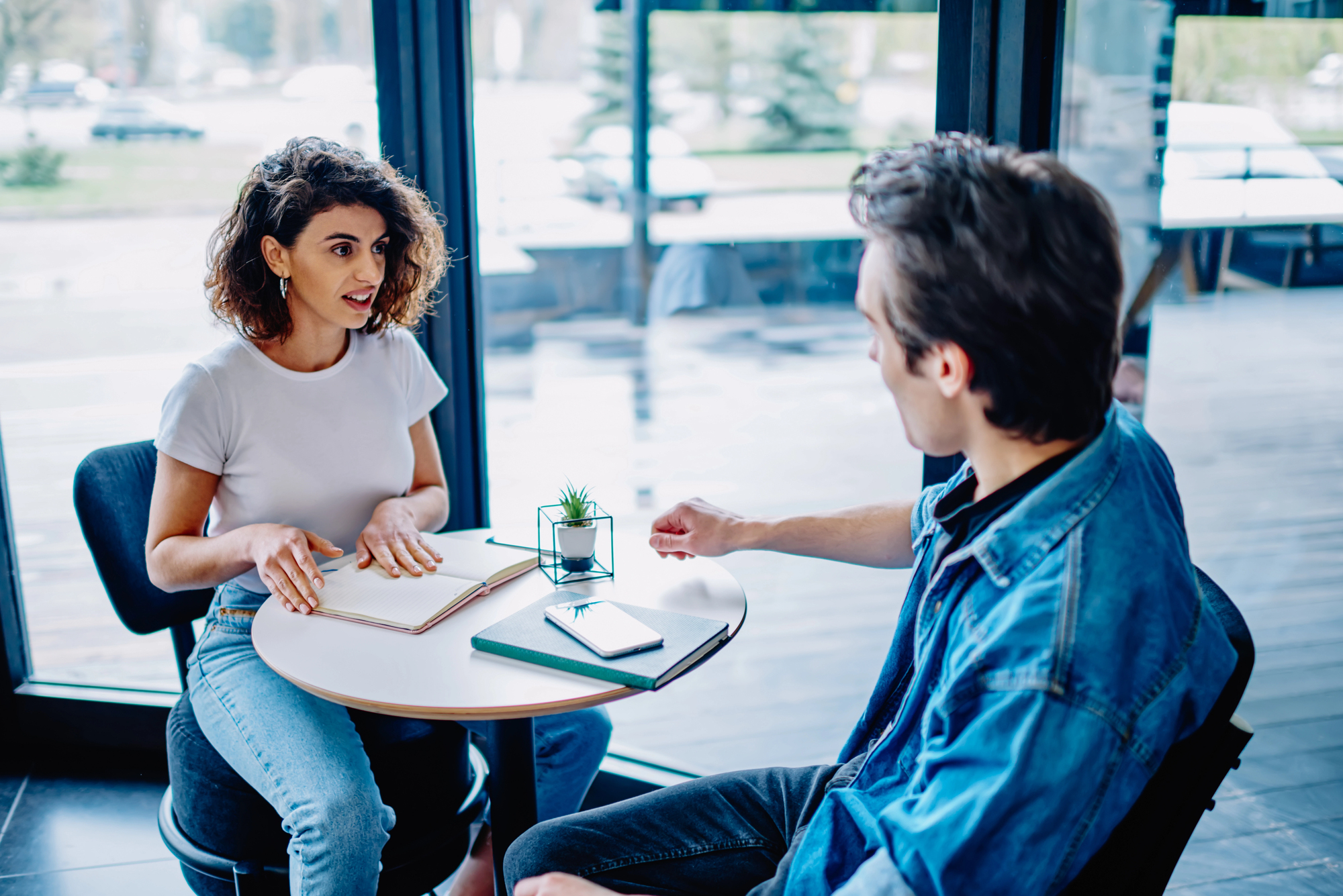 Two people sit at a round table by large windows. One has short curly hair and a white shirt, while the other has short hair and a denim jacket. They converse, with notebooks and a smartphone on the table. A small plant is between them.