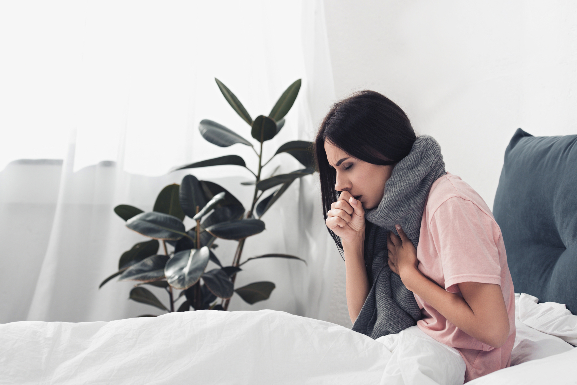 A woman sitting in bed with a blanket wrapped around her shoulders, coughing into her hand. A plant is visible in the background.