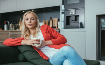 A person with long blonde hair, wearing a red cardigan and blue jeans, sits on a green couch in a modern kitchen holding a mug. The kitchen features sleek appliances and wooden countertops.