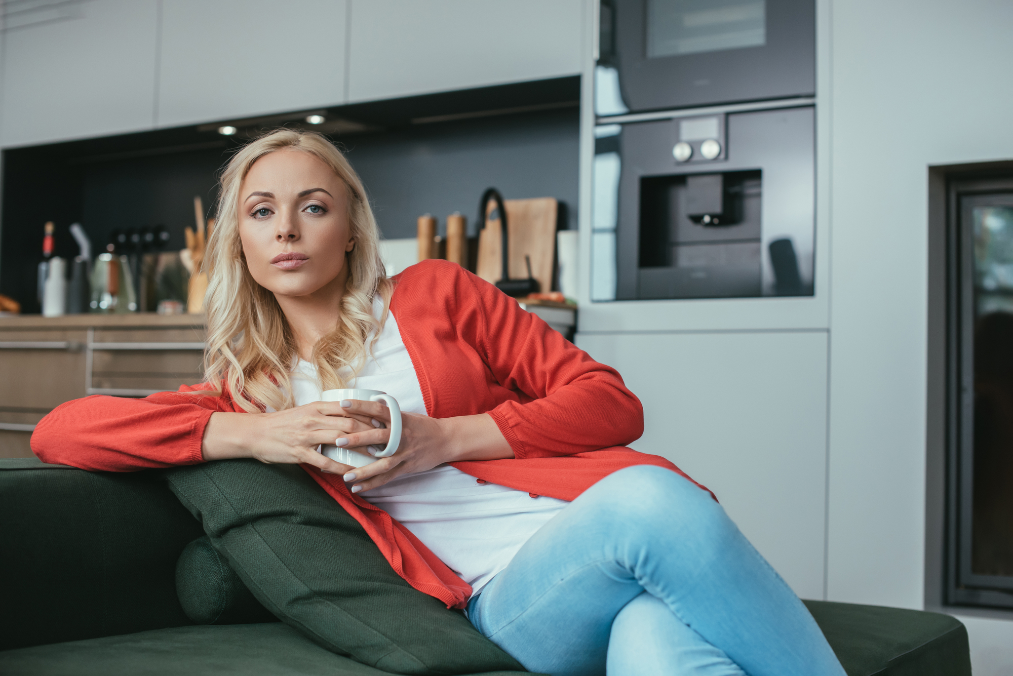 A person with long blonde hair, wearing a red cardigan and blue jeans, sits on a green couch in a modern kitchen holding a mug. The kitchen features sleek appliances and wooden countertops.