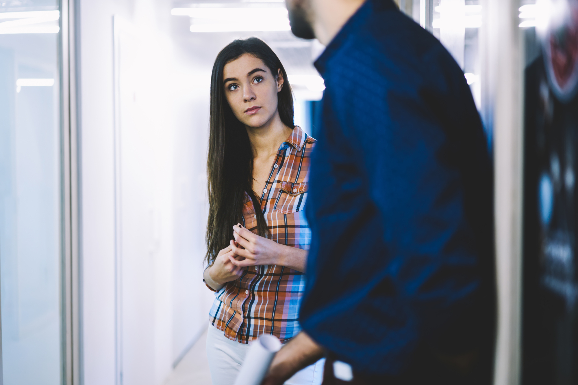 A woman in a plaid shirt stands in a hallway, looking at a man facing her. The man is wearing a blue shirt and holding a rolled-up paper. The scene suggests they are engaged in a conversation or meeting. The hallway is brightly lit.