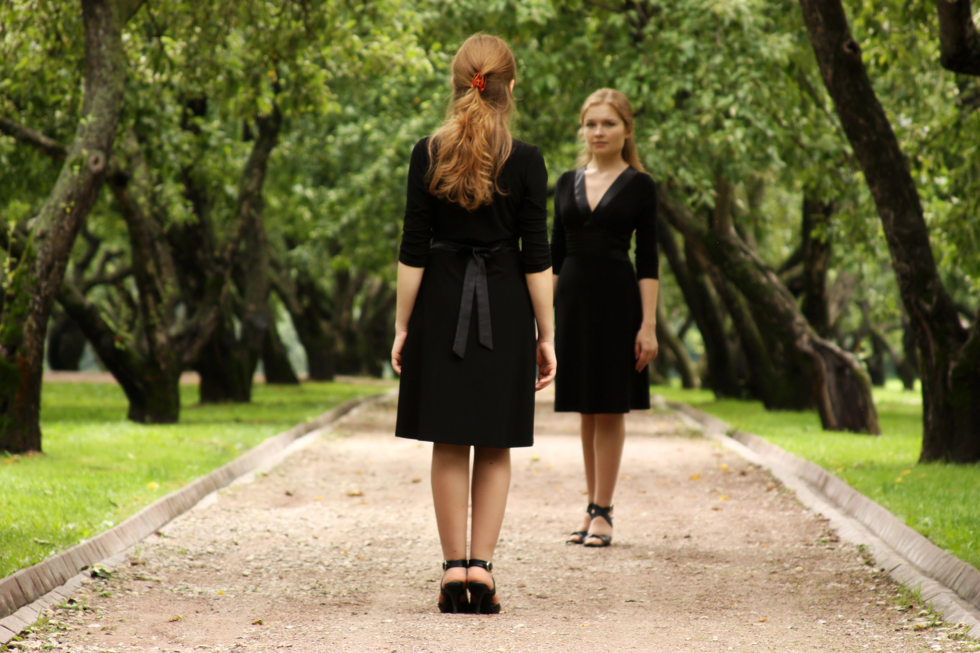 Two women in black dresses stand facing each other on a tree-lined path in a park. One woman has her back to the camera, showcasing the lush green surroundings.