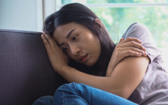 A woman with long dark hair sits on a couch, leaning her head against the cushion. She looks pensive and wraps her arms around herself, with a window showing greenery in the background.