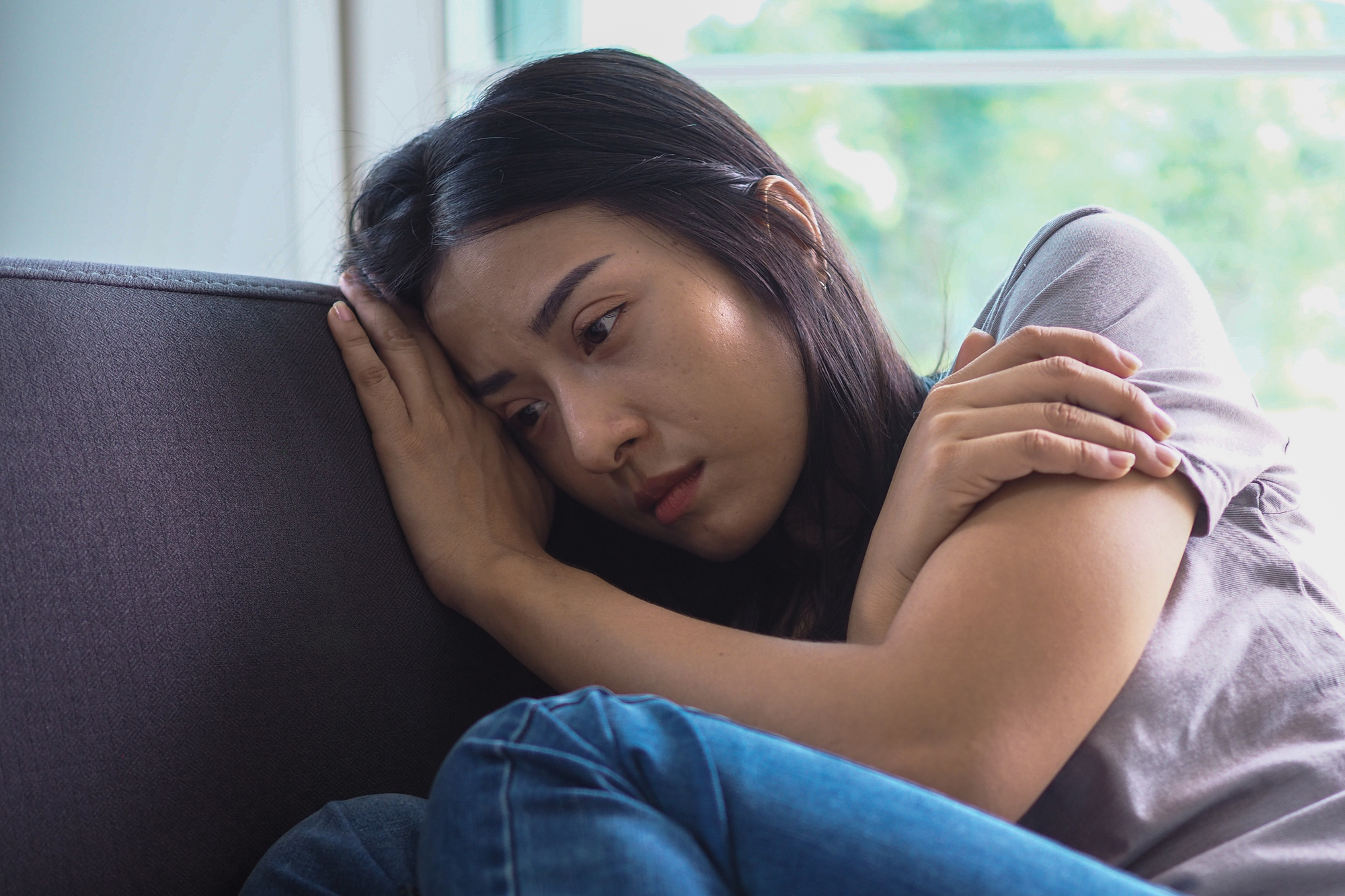 A woman with long dark hair sits on a couch, leaning her head against the cushion. She looks pensive and wraps her arms around herself, with a window showing greenery in the background.