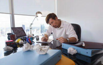 A man sits at a cluttered office desk, focused on writing. Crumpled paper balls surround him, and various folders and office supplies are scattered on the glass surface. A large window offers a view of a city skyline in the background.