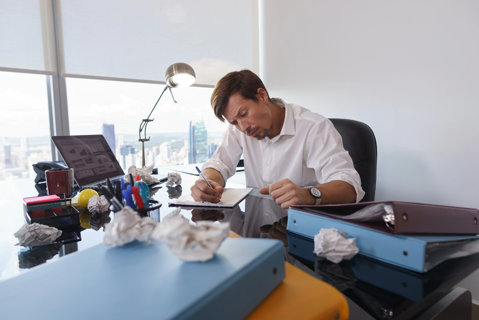 A man sits at a cluttered office desk, focused on writing. Crumpled paper balls surround him, and various folders and office supplies are scattered on the glass surface. A large window offers a view of a city skyline in the background.