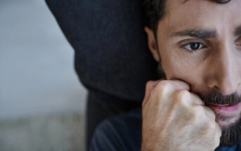 Close-up of a person with a beard, resting their head on a dark surface. Their hand is clenched near their face, and they appear deep in thought. The background is blurred.