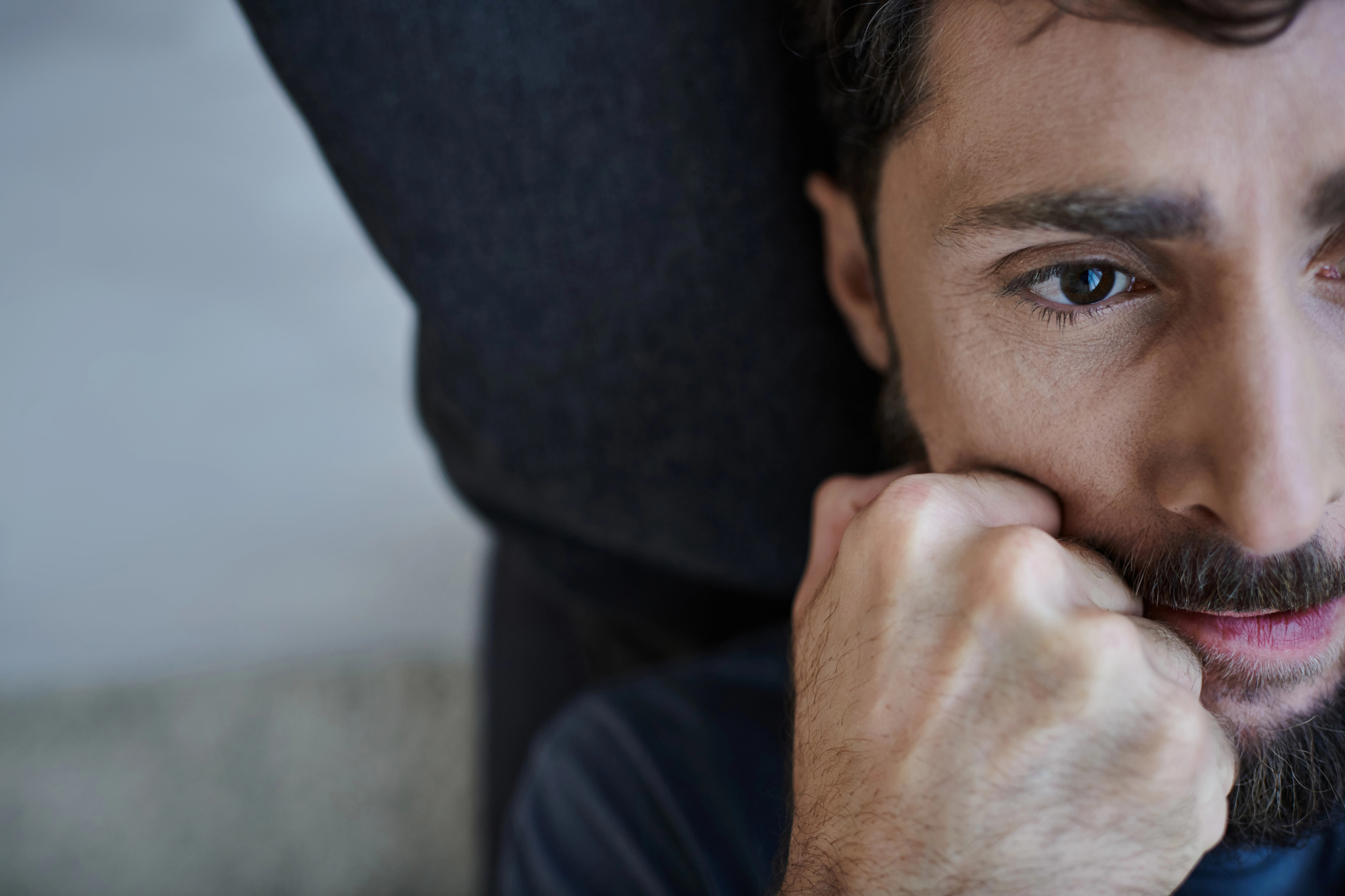 Close-up of a person with a beard, resting their head on a dark surface. Their hand is clenched near their face, and they appear deep in thought. The background is blurred.