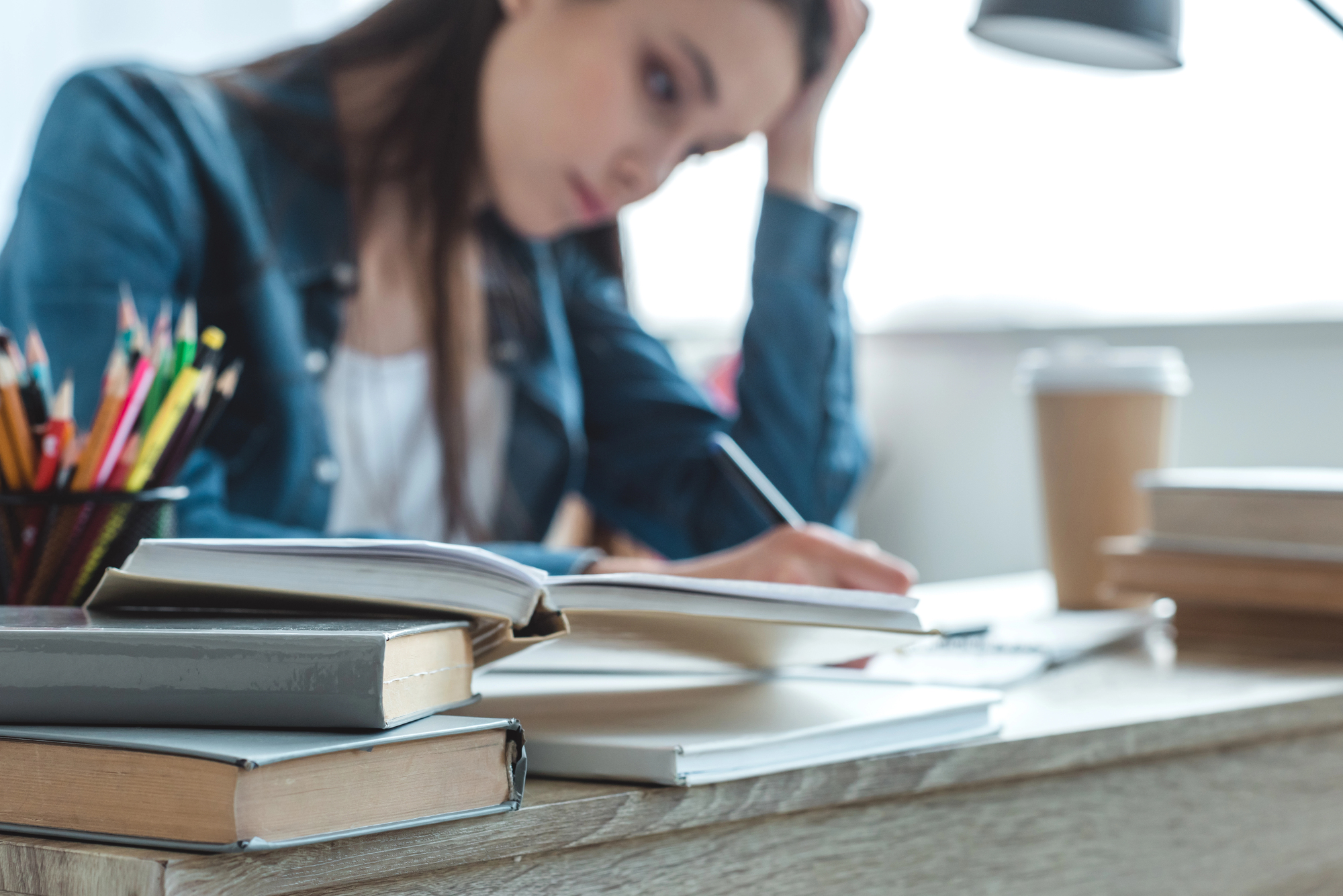 A woman with long brown hair sits at a desk, focused on writing in a notebook. Open books surround her, and a cup of coffee is nearby. She wears a denim jacket and leans her head on her hand, appearing deep in thought.