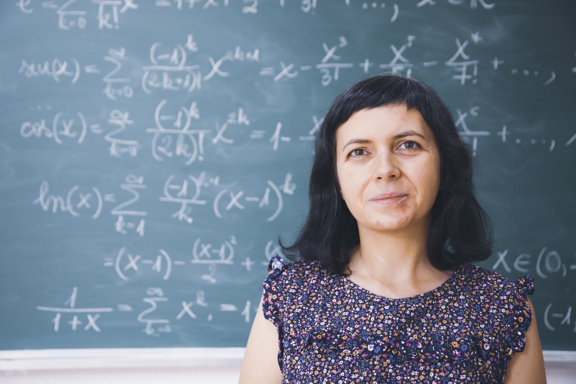 A woman with dark hair stands in front of a blackboard covered with mathematical equations. She is wearing a patterned sleeveless top and looks directly into the camera.