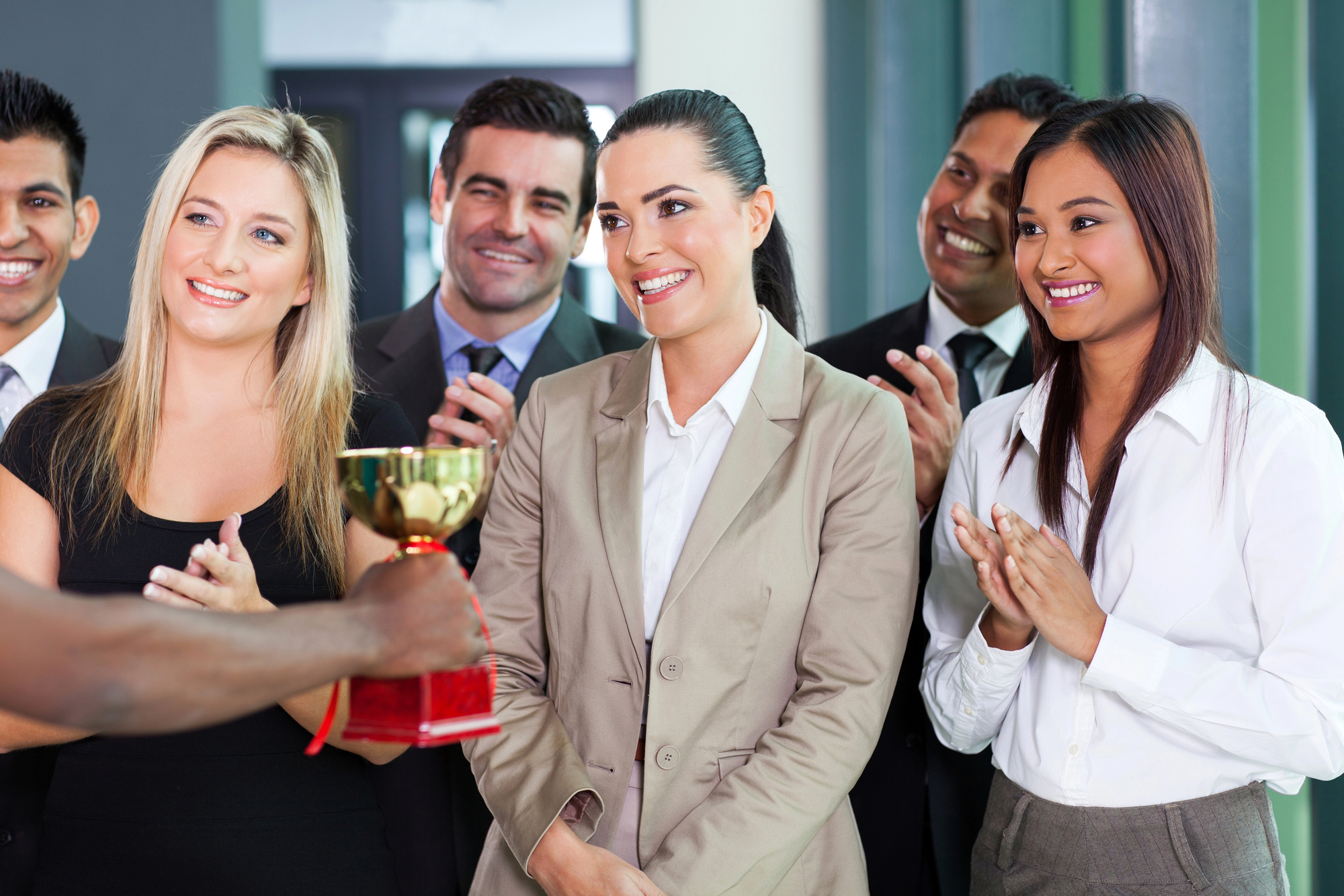 A group of diverse professionals standing and clapping as a person receives a trophy. The individuals are dressed in business attire, showing expressions of happiness and encouragement in an office setting.