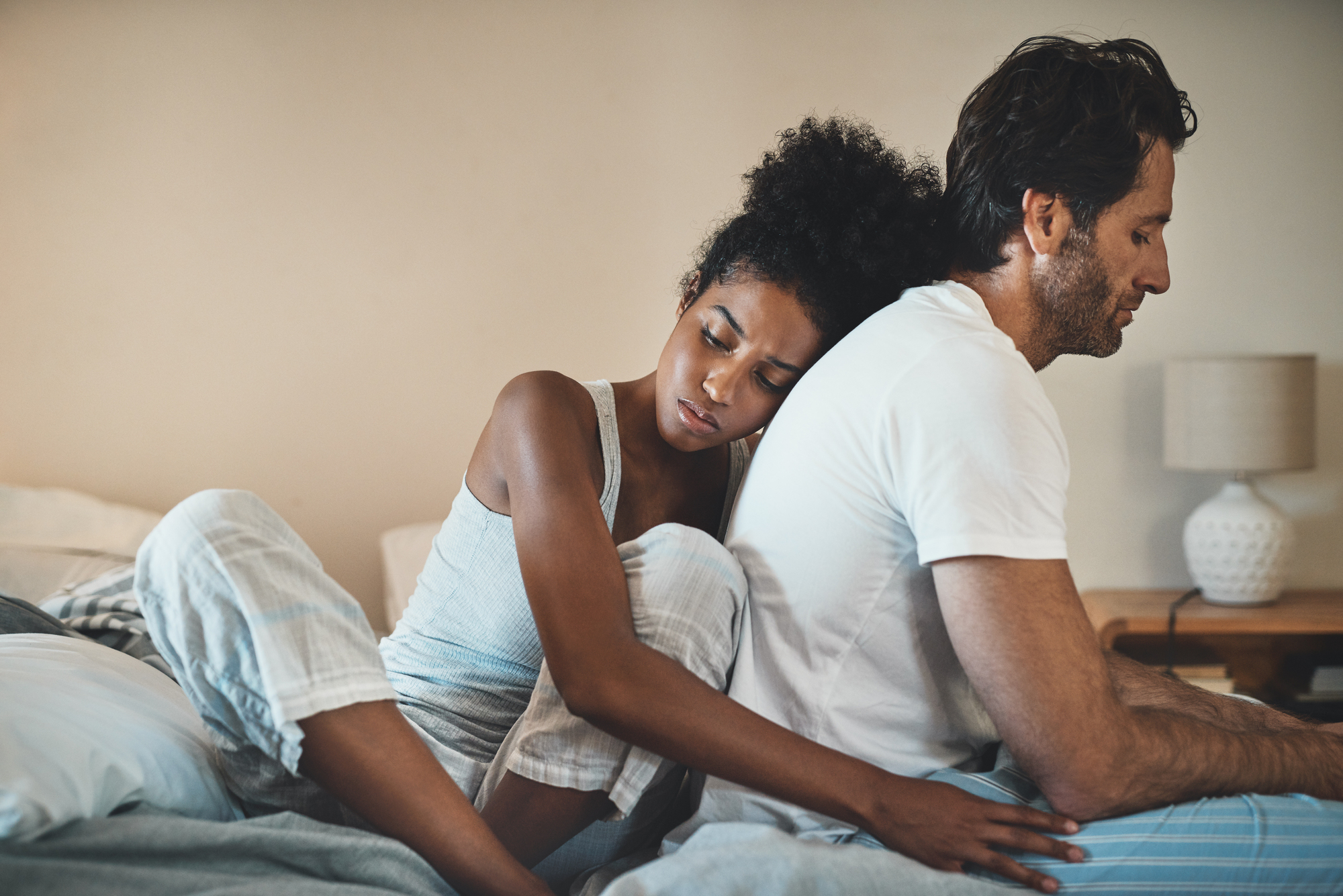 A woman in pajamas rests her head on a man's back as they sit on a bed. The room is softly lit, with a lamp and a wooden nightstand in the background. Both appear contemplative and relaxed.