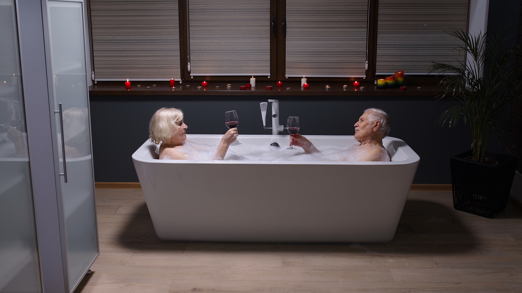 An elderly couple relaxes in a large bathtub filled with bubbles, holding glasses of red wine. The room is dimly lit with candles, and the window blinds are closed. A potted plant is visible on the right side of the room.