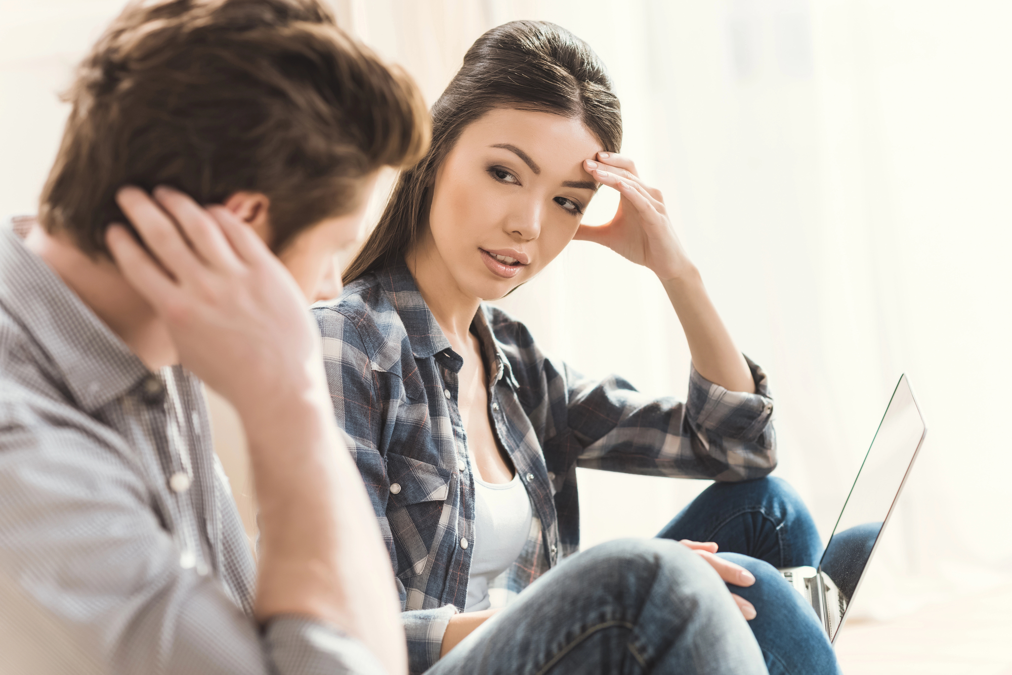 A woman and man are sitting together indoors, engaged in a serious conversation. The woman, wearing a plaid shirt, looks at the man with a concerned expression. A laptop rests on her lap. The setting appears comfortable and well-lit.