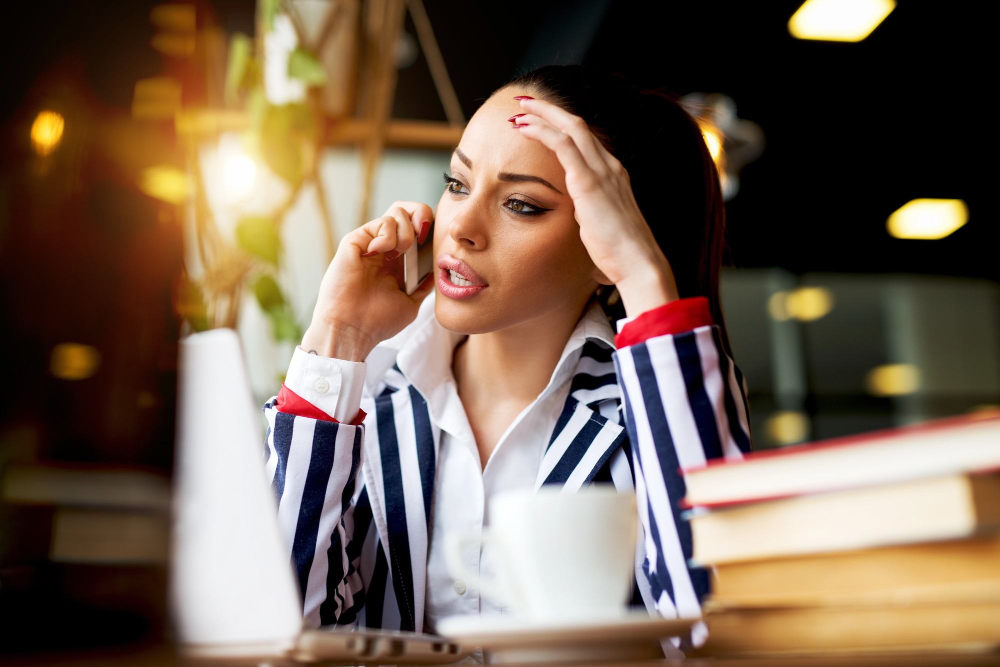 A woman in a striped blazer is sitting at a table, speaking on her phone. She looks concerned, with one hand on her head. In front of her are a laptop, books, and a cup, and there's warm lighting in the background.