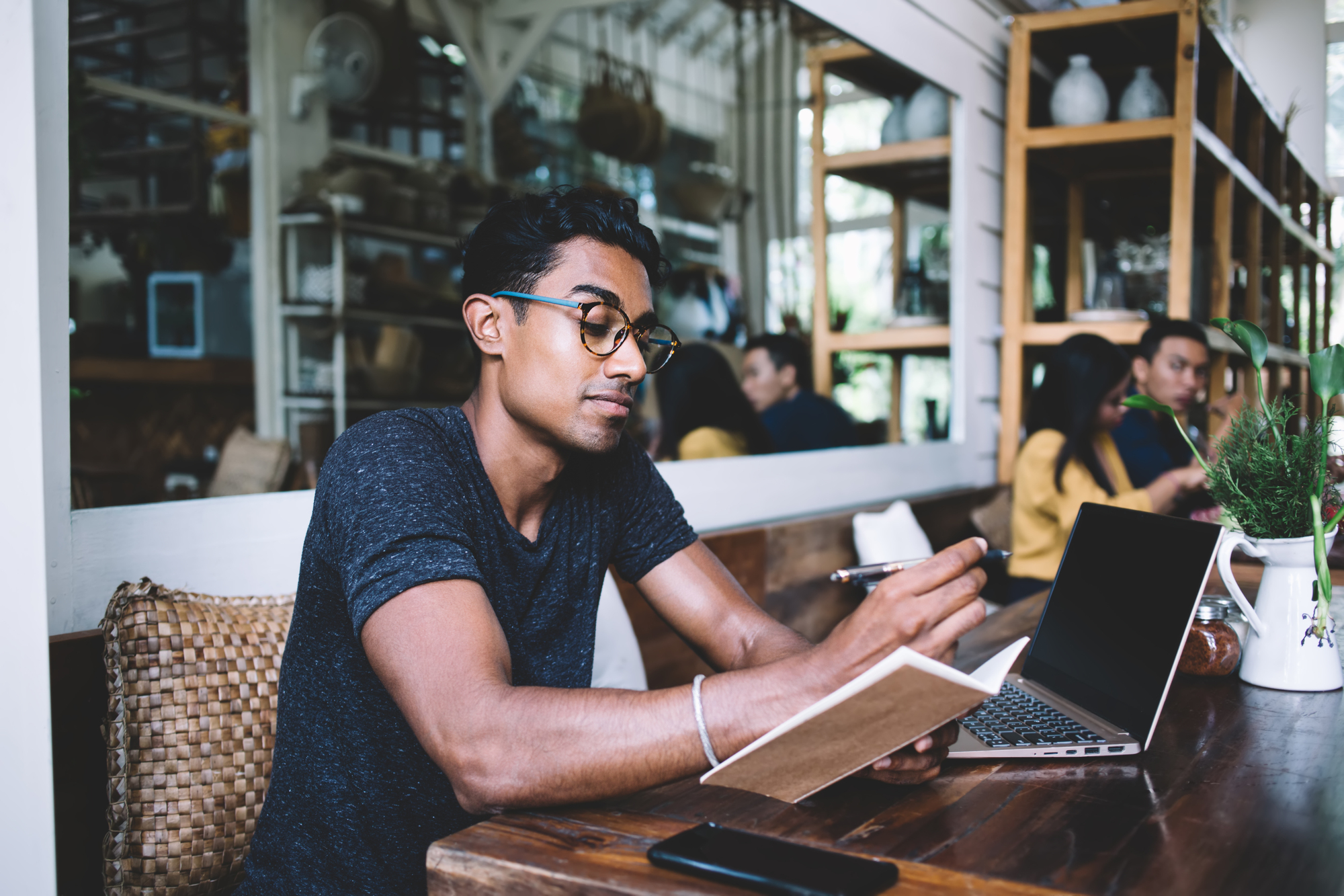 A man wearing glasses is sitting at a wooden table in a café, holding a notebook and pen. A laptop and smartphone are on the table. Shelves and other patrons are visible in the background.