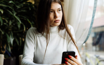 A woman with long brown hair in a white turtleneck is sitting by a window, holding a smartphone. She gazes to the side, appearing thoughtful. Plants and soft curtains are in the background, and natural light streams in through the window.