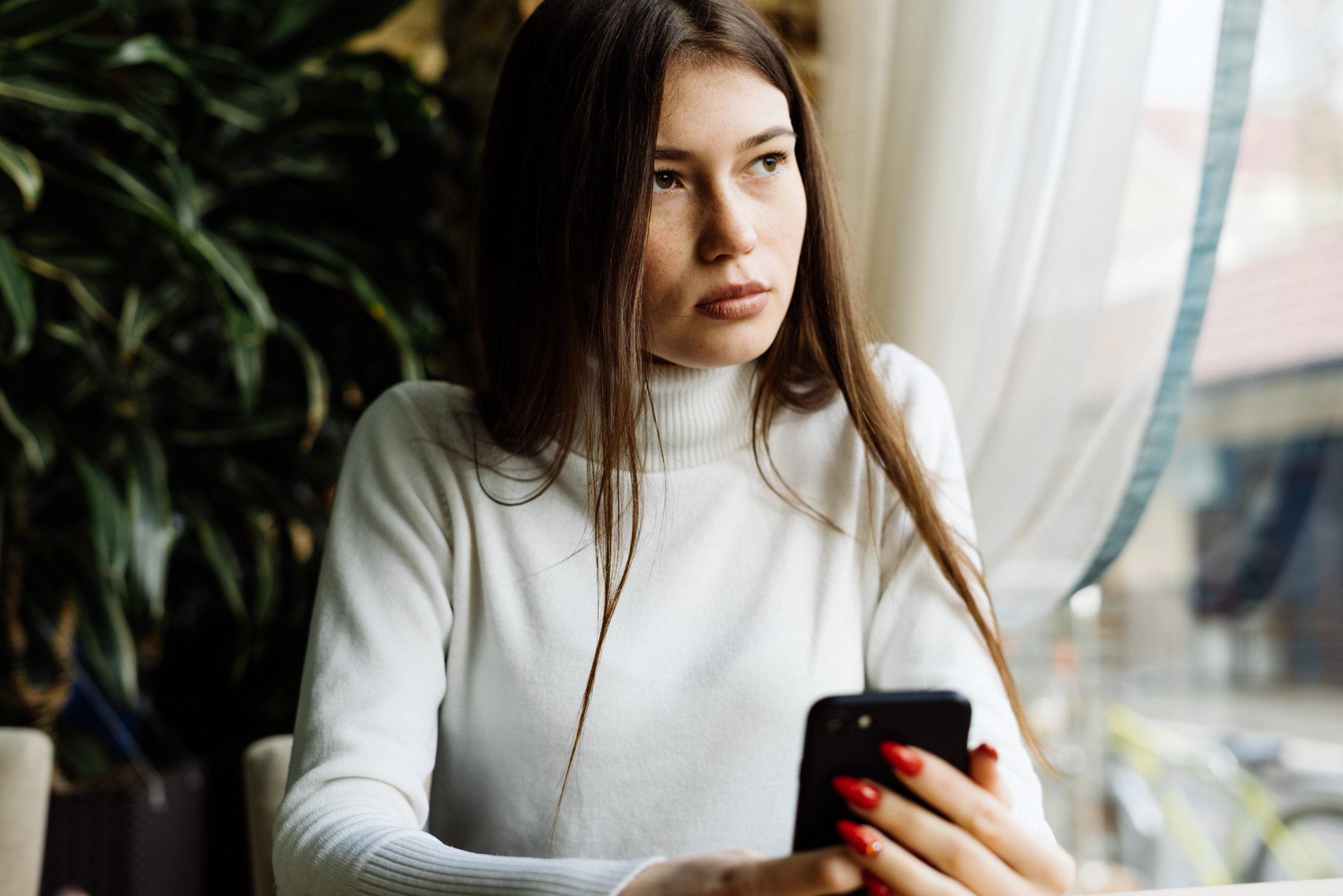 A woman with long brown hair in a white turtleneck is sitting by a window, holding a smartphone. She gazes to the side, appearing thoughtful. Plants and soft curtains are in the background, and natural light streams in through the window.