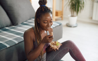 A woman with braided hair sits on the floor in a living room, eating granola with sliced apples from a bowl. She is smiling and wearing athletic wear, with a couch and a potted plant in the background.