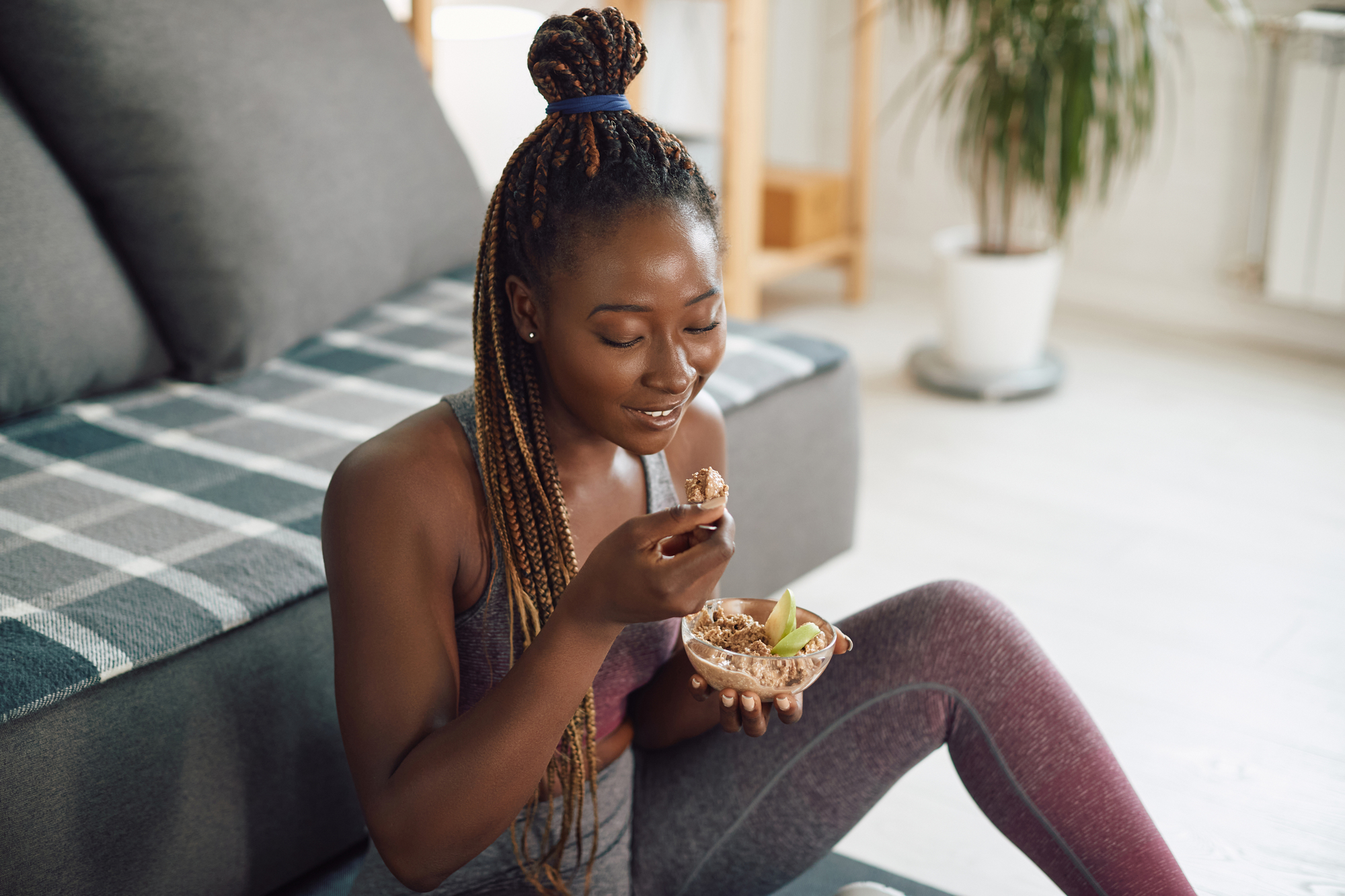 A woman with braided hair sits on the floor in a living room, eating granola with sliced apples from a bowl. She is smiling and wearing athletic wear, with a couch and a potted plant in the background.