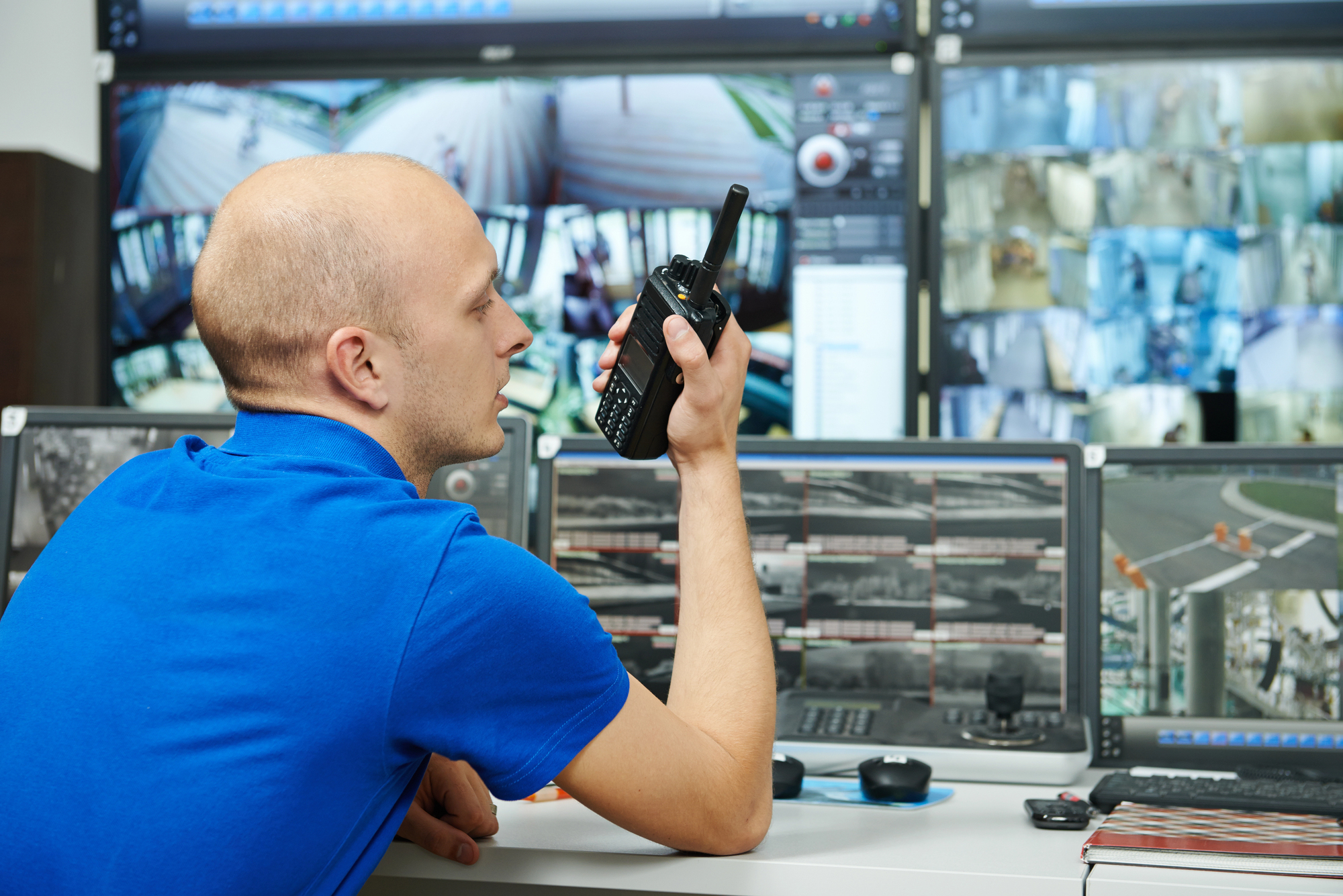 A man in a blue shirt holds a walkie-talkie while sitting at a desk filled with monitors displaying security footage. He appears to be communicating or coordinating based on the display images.