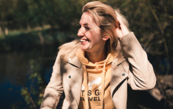 A person wearing a beige coat and yellow hoodie smiles while brushing back their hair. They are outdoors, with blurred greenery in the background, enjoying a sunny day.