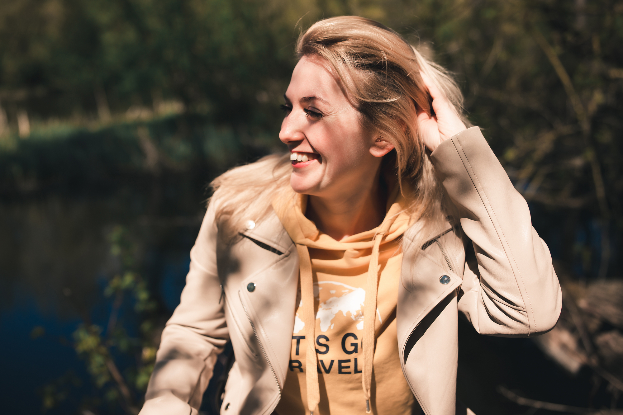 A person wearing a beige coat and yellow hoodie smiles while brushing back their hair. They are outdoors, with blurred greenery in the background, enjoying a sunny day.