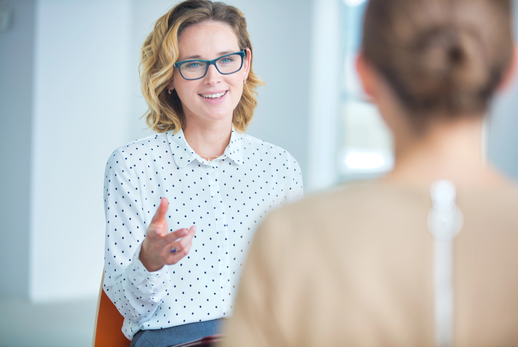 A woman wearing glasses and a white polka dot blouse is sitting and talking to another person whose back is to the camera. She appears engaged in the conversation, gesturing with her hand, in a bright, modern room.