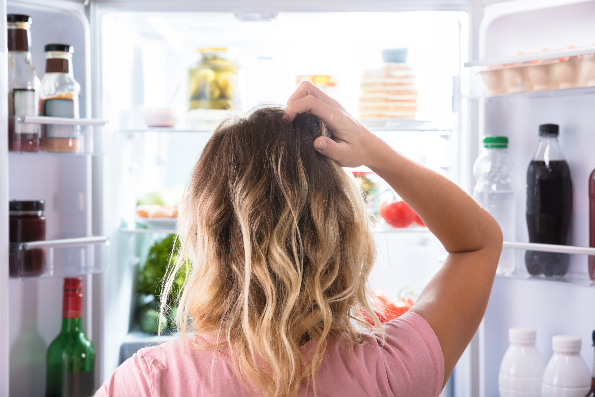 A person with wavy blonde hair stands in front of an open refrigerator, scratching their head thoughtfully. The fridge is filled with various food items, including vegetables, drinks, and condiments.