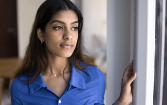 A woman with long hair stands by a window, wearing a blue shirt. She gazes outside with a thoughtful expression. The background is softly blurred, suggesting an indoor setting.