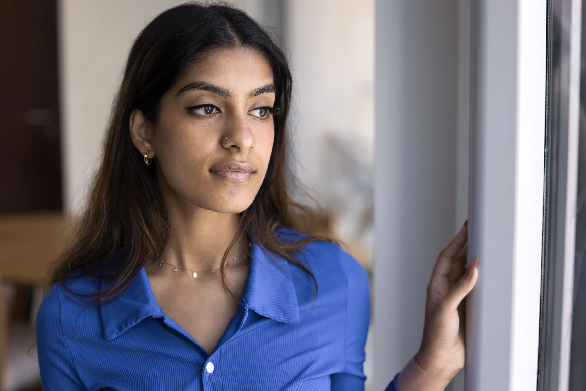 A woman with long hair stands by a window, wearing a blue shirt. She gazes outside with a thoughtful expression. The background is softly blurred, suggesting an indoor setting.