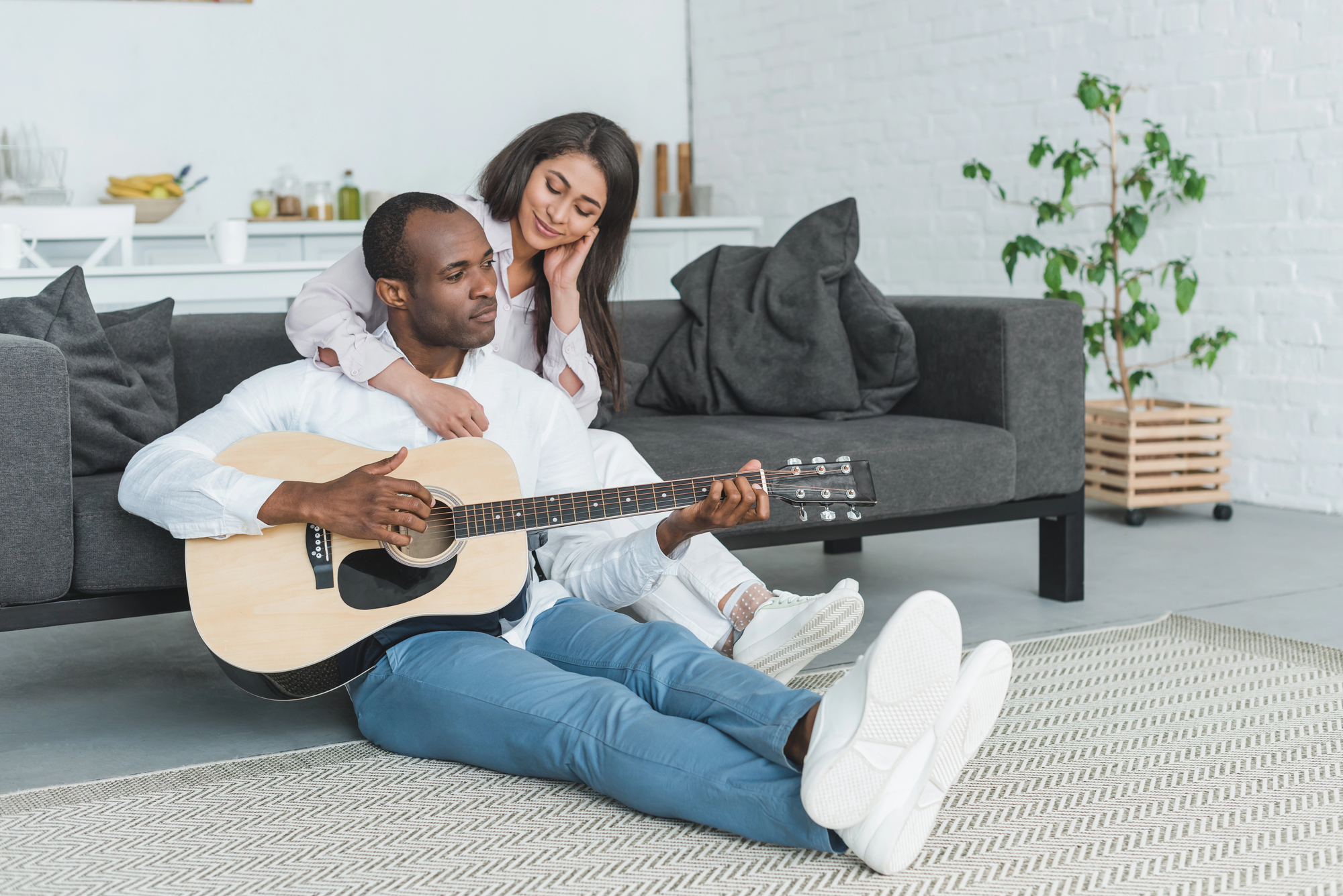 A man sitting on the floor plays an acoustic guitar while a woman rests her chin on his shoulder, listening contently. They are in a bright living room with a sofa, potted plant, and decorative items in the background.