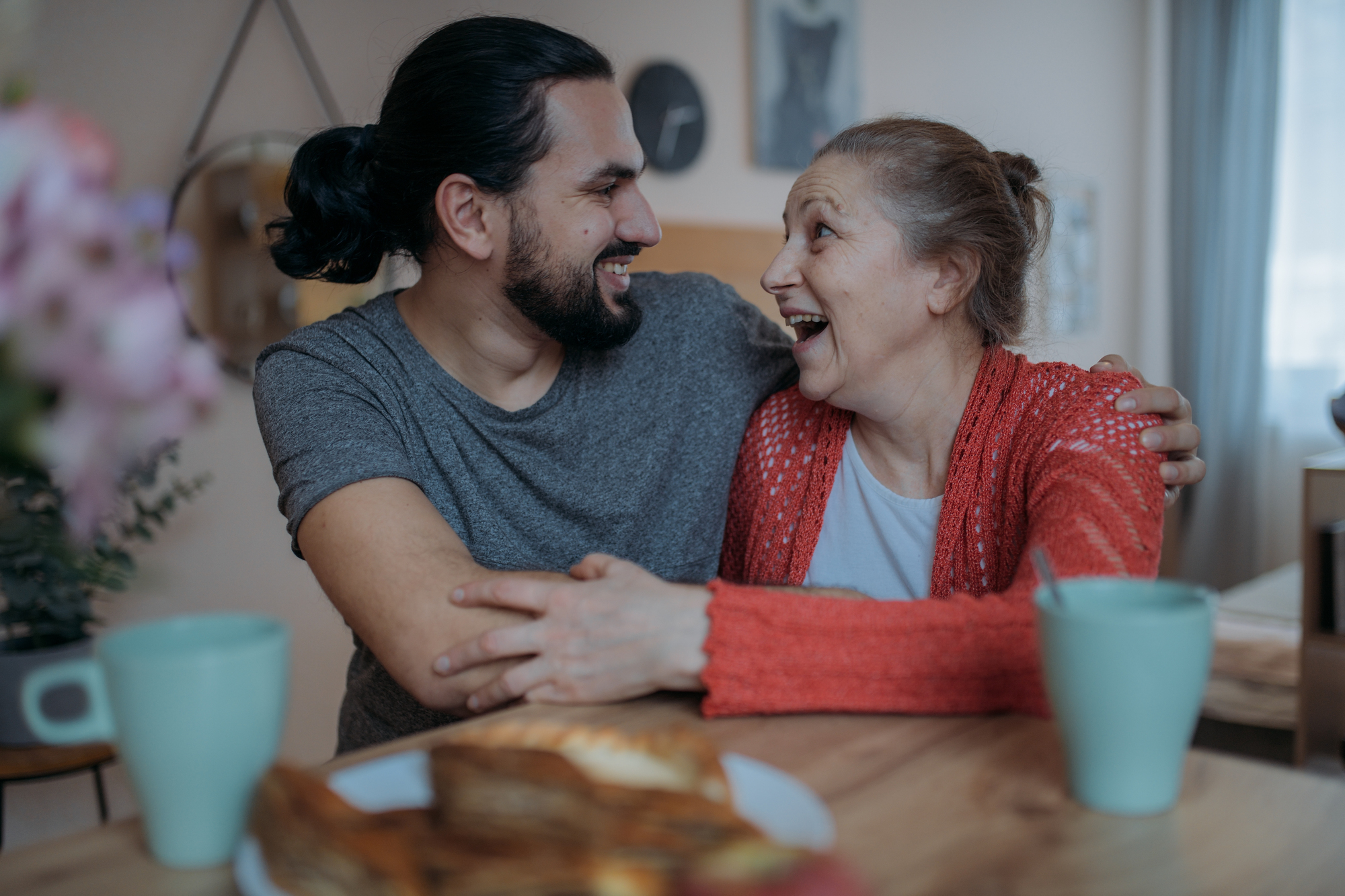 A smiling man and woman sit at a table, embracing. They appear to share a joyful moment, surrounded by coffee mugs and pastries. The room has a cozy and relaxed atmosphere.