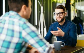 Two men are sitting at a table in a modern cafe. One is wearing glasses and a blue denim shirt, gesturing while talking. The other, in a plaid shirt, is listening. A coffee mug is placed on the table in front of them.