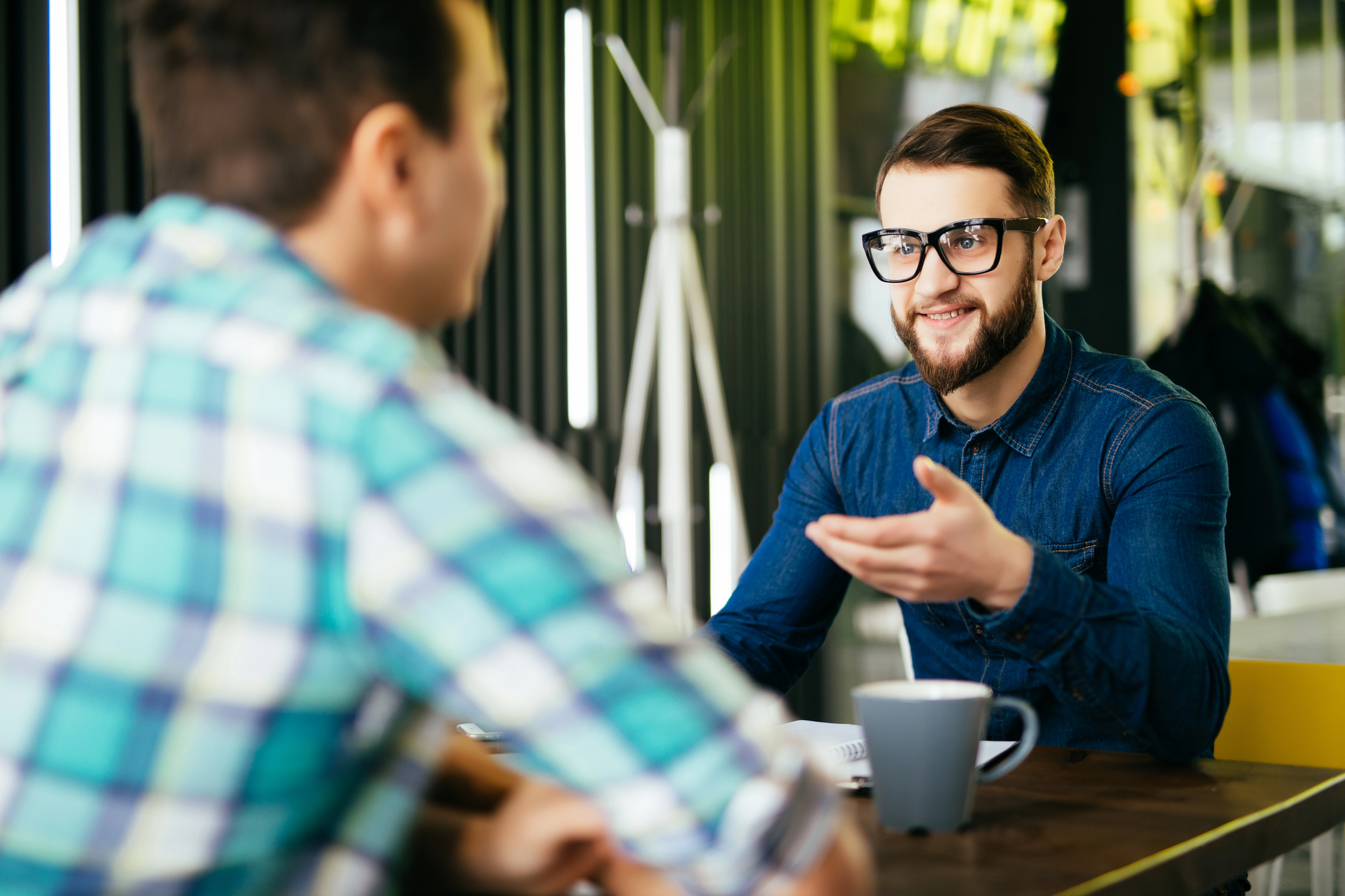 Two men are sitting at a table in a modern cafe. One is wearing glasses and a blue denim shirt, gesturing while talking. The other, in a plaid shirt, is listening. A coffee mug is placed on the table in front of them.