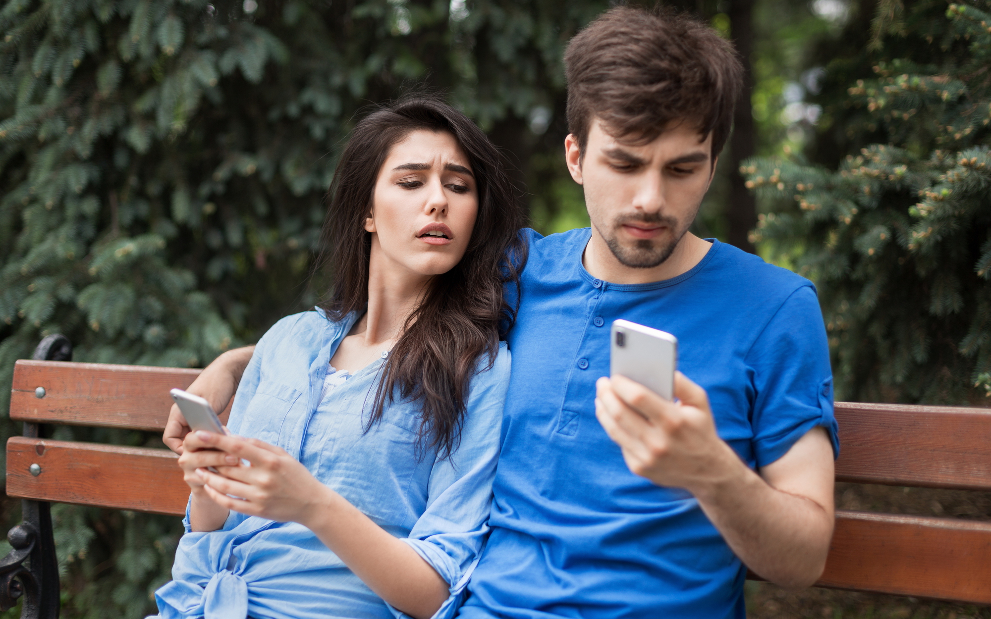 A woman and man sitting on a bench. The woman looks at the man's phone with a curious expression. Both are wearing blue tops, and they are surrounded by lush green foliage.