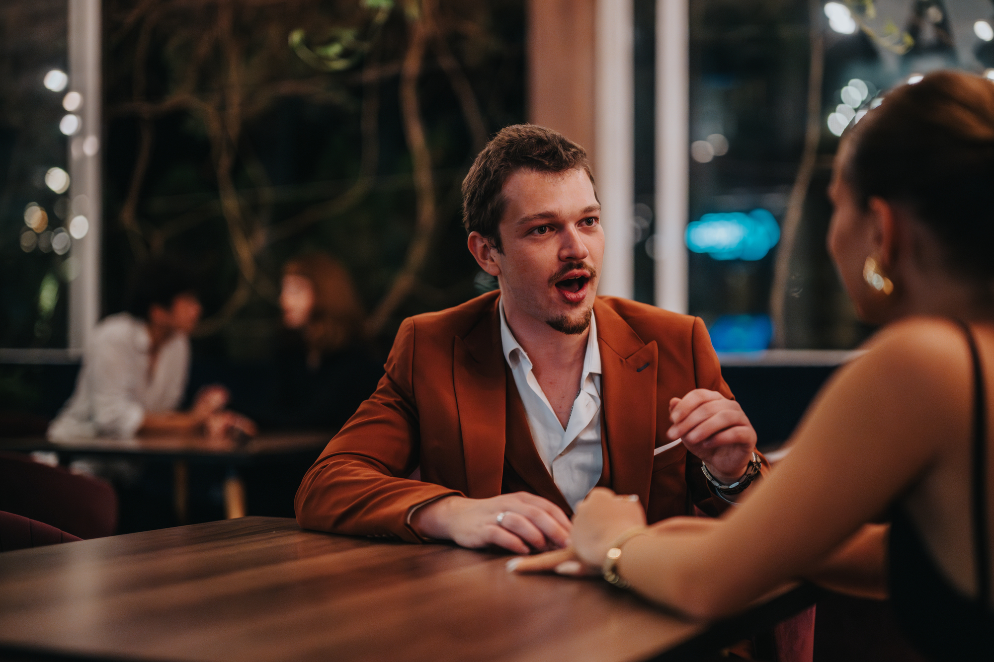 A man in a brown blazer is talking animatedly to a woman across a table in a dimly lit café. The woman is wearing a black top, and there are other people in the background. The atmosphere is cozy and intimate.