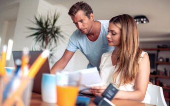 A man and woman are working together at a desk, reviewing paperwork. The man stands leaning over a laptop, while the woman sits, holding documents. The desk holds office supplies and a glass of orange juice. A plant is visible in the background.