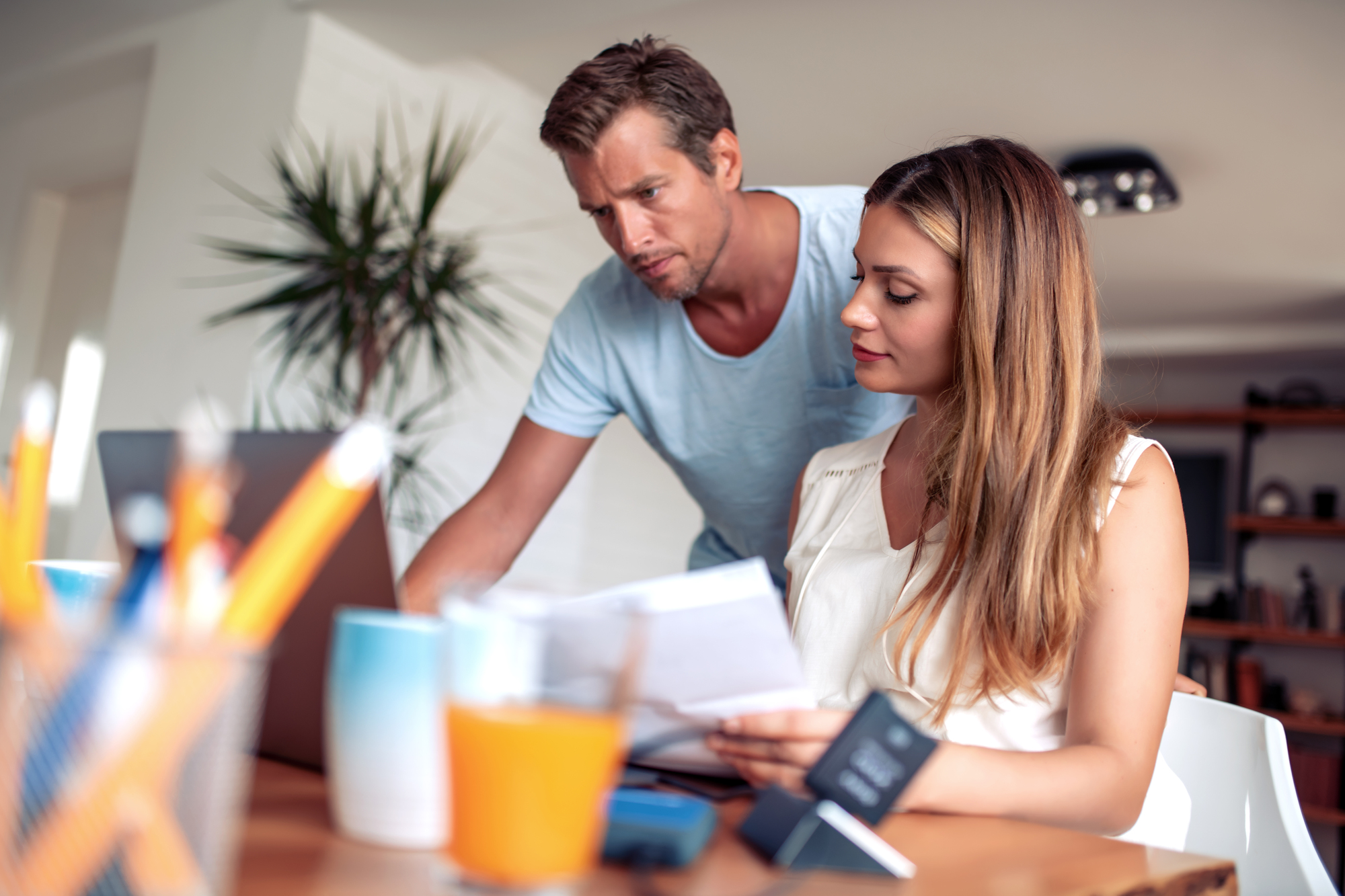 A man and woman are working together at a desk, reviewing paperwork. The man stands leaning over a laptop, while the woman sits, holding documents. The desk holds office supplies and a glass of orange juice. A plant is visible in the background.