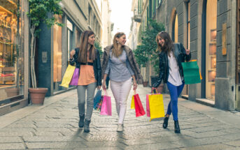 Three women walking on a city street, joyfully carrying colorful shopping bags. They are dressed in casual clothes and chatting, with shops and greenery visible in the background.