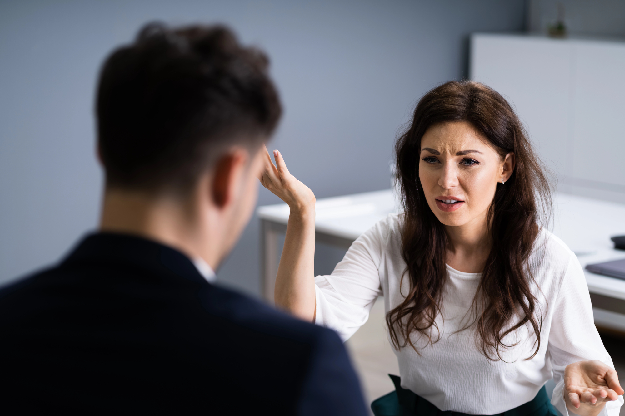 A woman with long hair, wearing a white shirt, is sitting in an office and appears to be engaged in a serious conversation with a man in a suit, whose back is to the camera. She is gesturing with her hands and has a concerned expression.