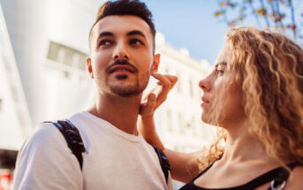 A woman with curly hair gently touches the face of a man with short dark hair and facial hair. They are outdoors, with a modern building and trees in the background. Both are casually dressed, and the atmosphere is calm and sunny.