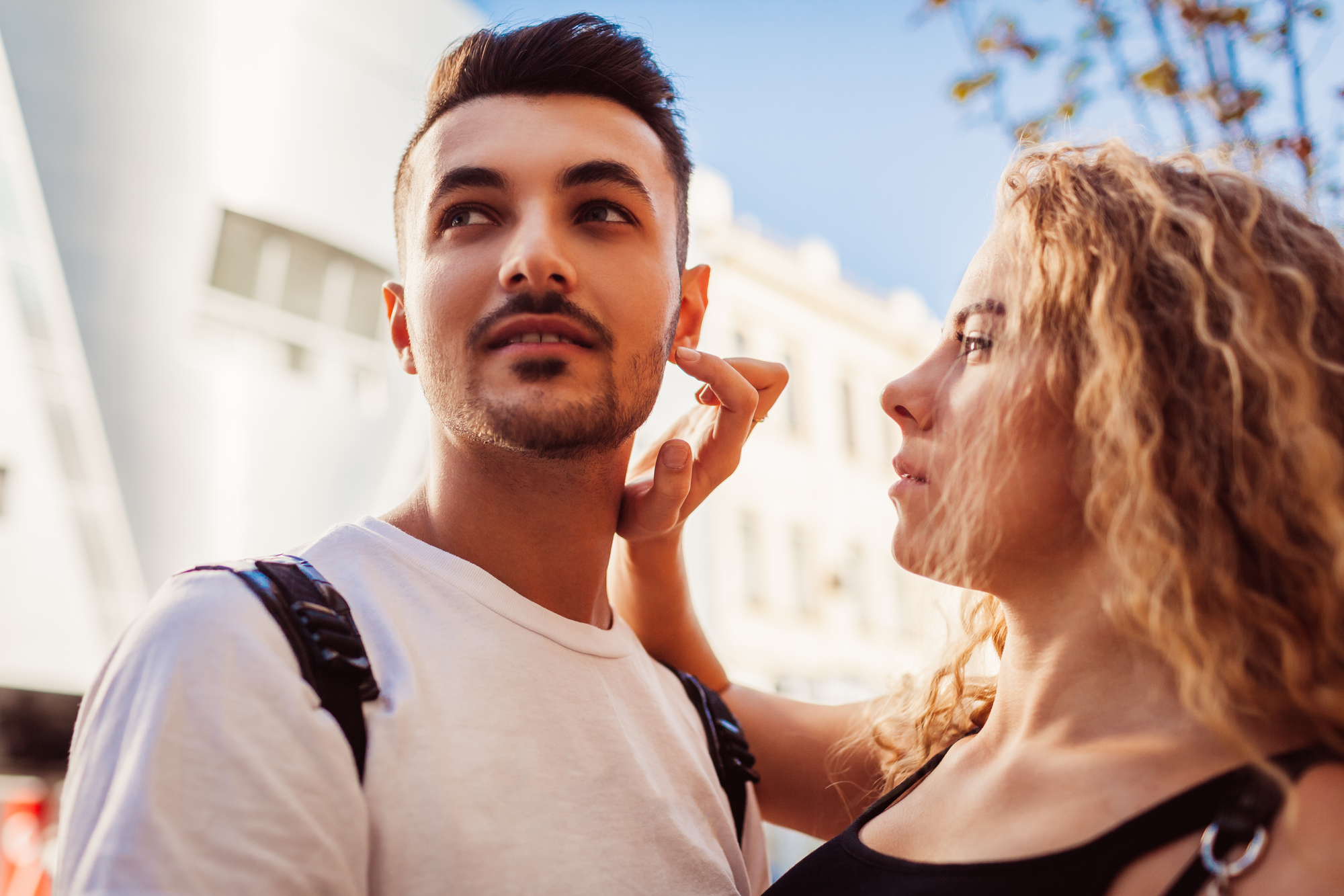 A woman with curly hair gently touches the face of a man with short dark hair and facial hair. They are outdoors, with a modern building and trees in the background. Both are casually dressed, and the atmosphere is calm and sunny.