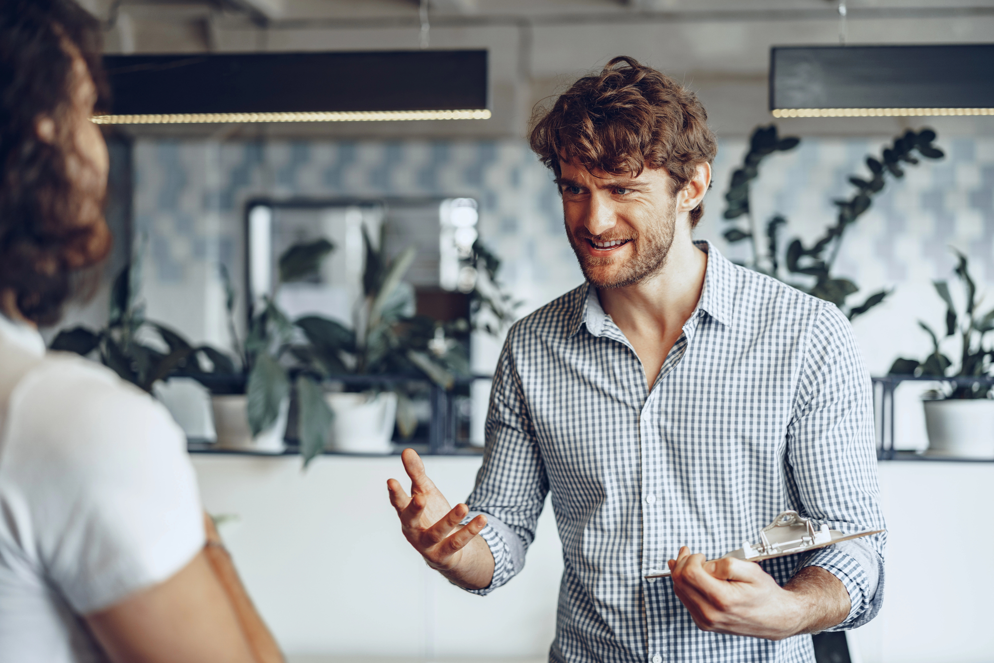 A man with curly hair and a checkered shirt is energetically speaking to another person indoors. He holds a tablet and gestures with his hands. The background features several potted plants on a shelf and soft lighting.