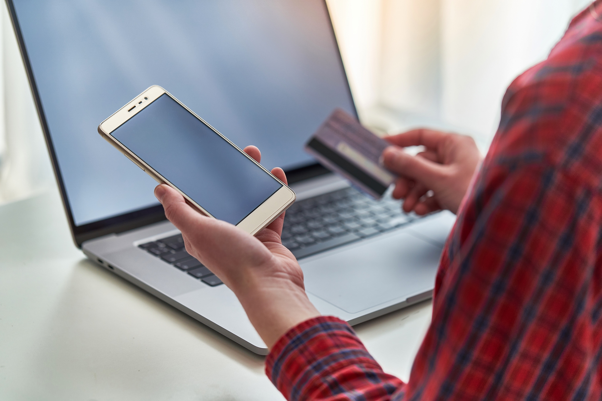 A person holding a smartphone in one hand and a credit card in the other, sitting in front of a laptop. They appear to be engaged in online shopping or a financial transaction. The person is wearing a red plaid shirt.