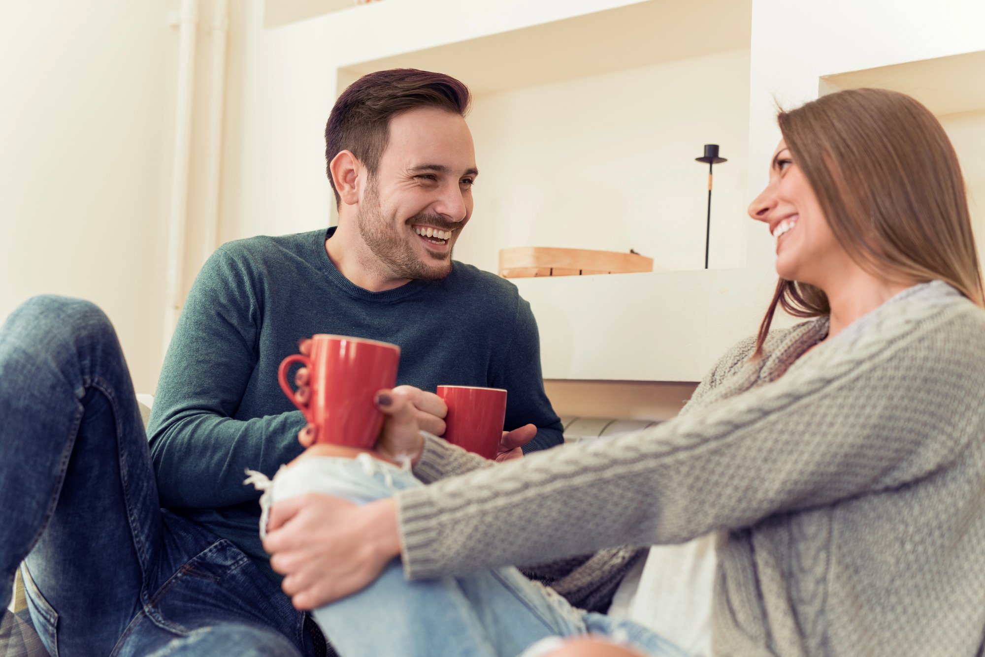 A man and a woman are sitting on the floor, smiling at each other while holding red mugs. They appear relaxed and happy, surrounded by a cozy, light-colored interior.