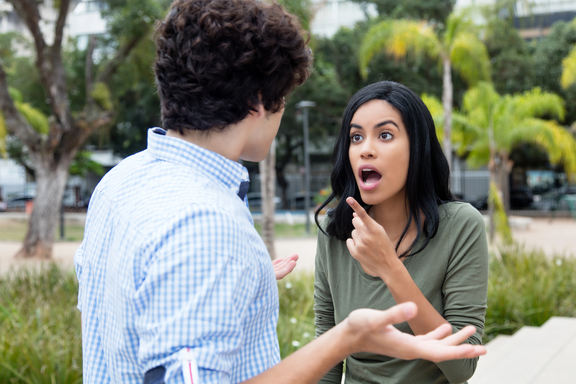 A man and woman stand outside in a park setting. The woman, wearing a green shirt, appears surprised or upset, pointing at the man. The man, in a blue checkered shirt, has his hands open in a questioning gesture. Trees and plants are in the background.