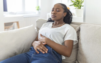 A woman sitting on a sofa holds her stomach with both hands, appearing uncomfortable. She is wearing a white shirt and blue jeans. In the background, there's a bright room with a window and a potted plant.